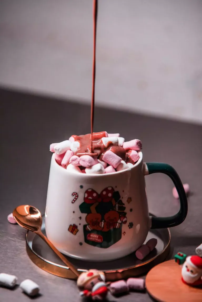 Taza decorada con una caja de regalos y galletas de navidad, en cuyo interior se ven nubes de gominola sobre las que cae un chorro de chocolate
