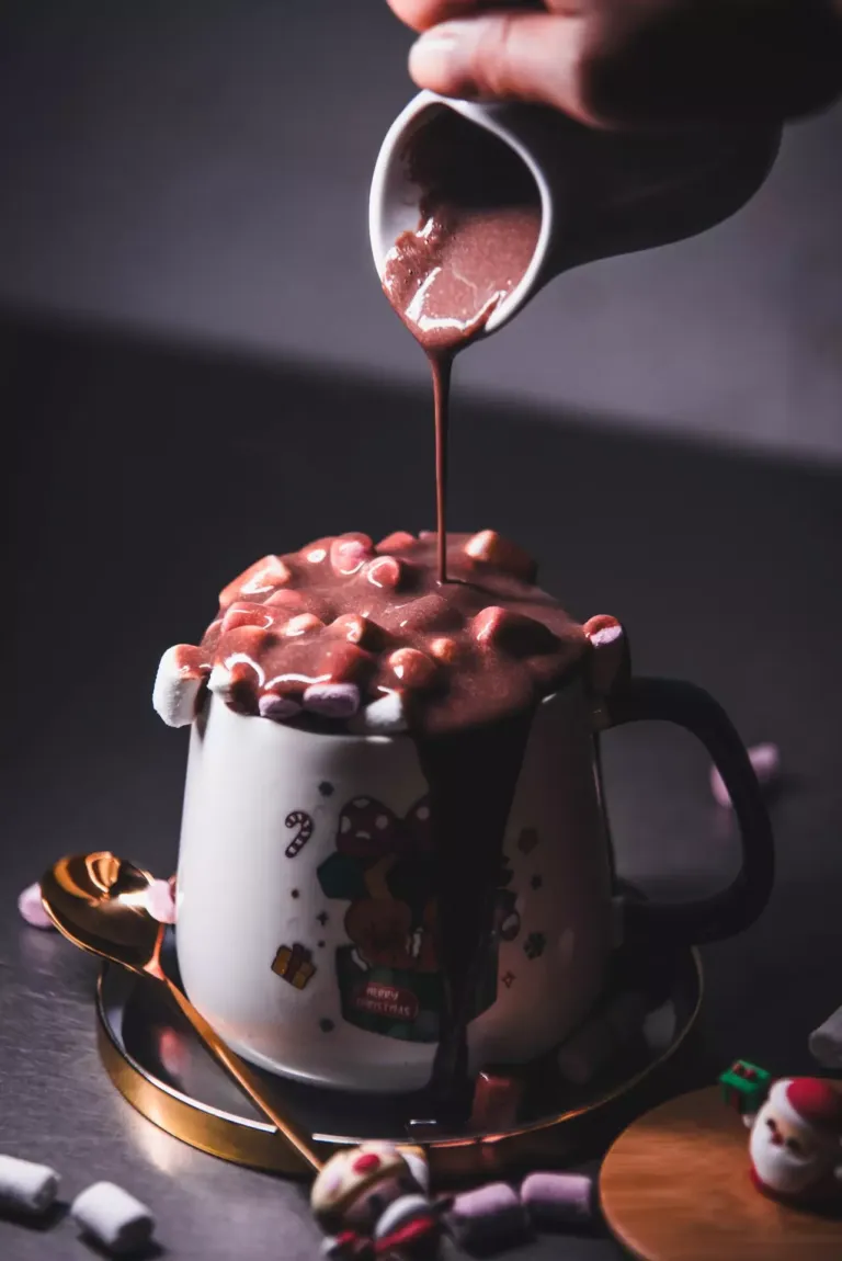Taza decorada con una caja de regalos y galletas de navidad, en cuyo interior se ven nubes de gominola sobre las que cae un chorro de chocolate